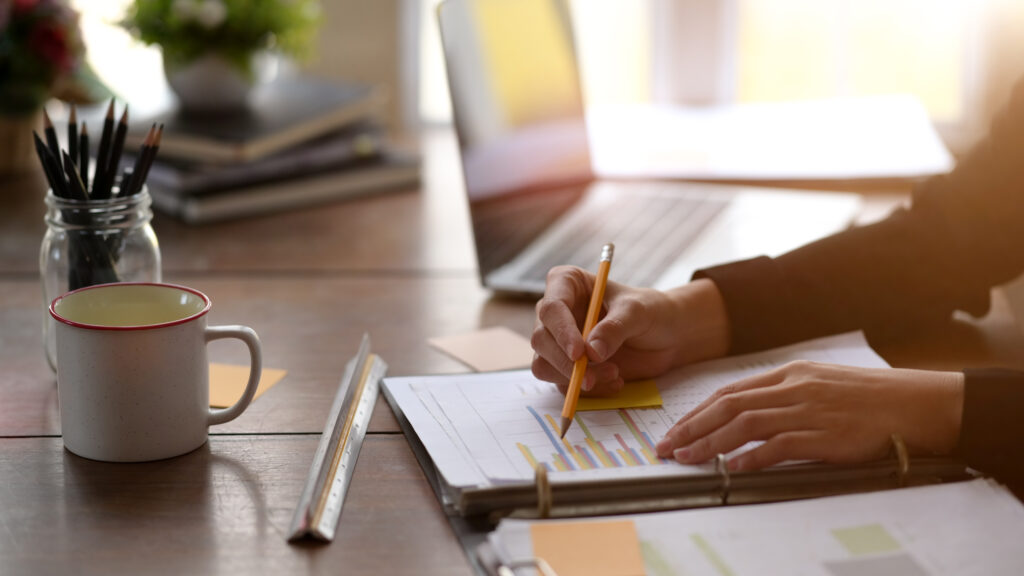 Table with a laptop and a mug. A person checking information on paper.